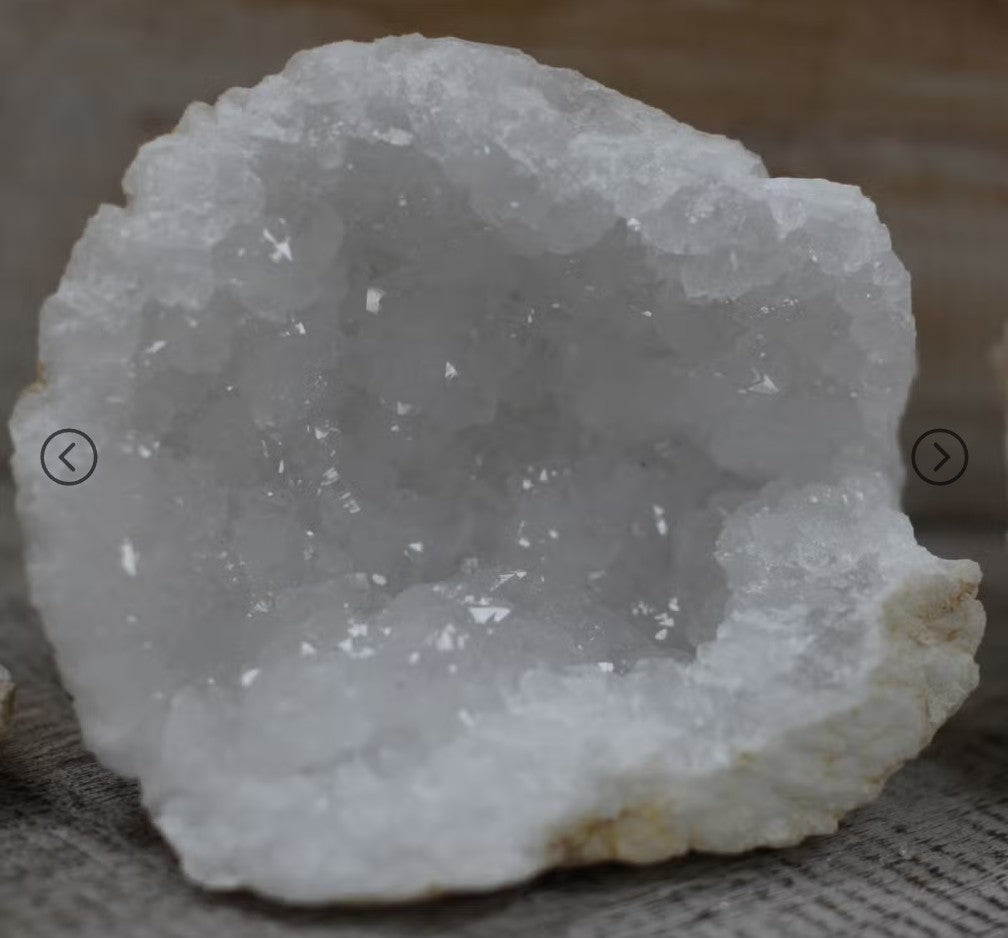 A white crystal geode dome with rough crystalline texture and a light beige outer matrix, photographed close-up on a neutral fabric surface.